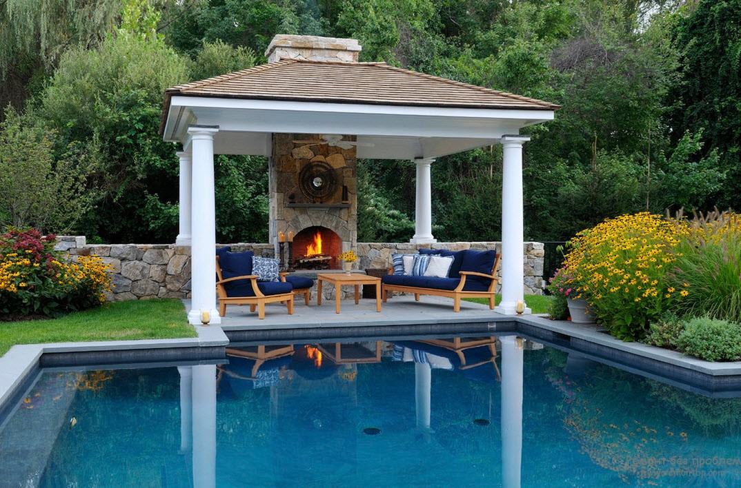 Een barbecuepaviljoen in klassieke stijl bevindt zich tegen de achtergrond van decoratieve bomen en bloemen, weerspiegeld in het oppervlak van het zwembadwater